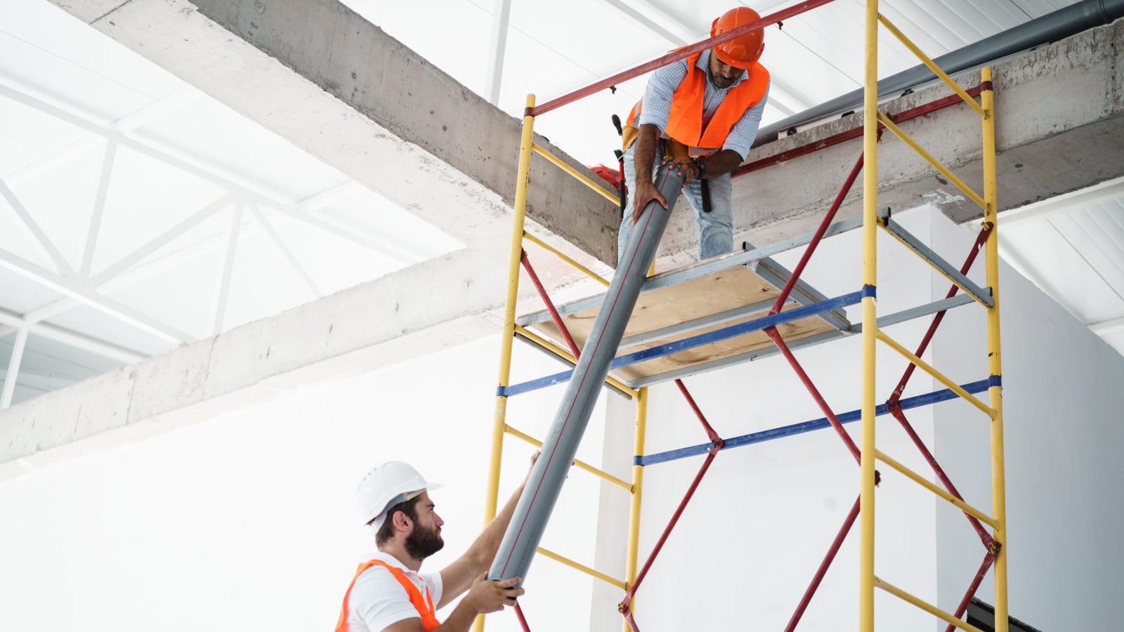 Two men builder in uniform drag the pipe on scaffolding at construction site