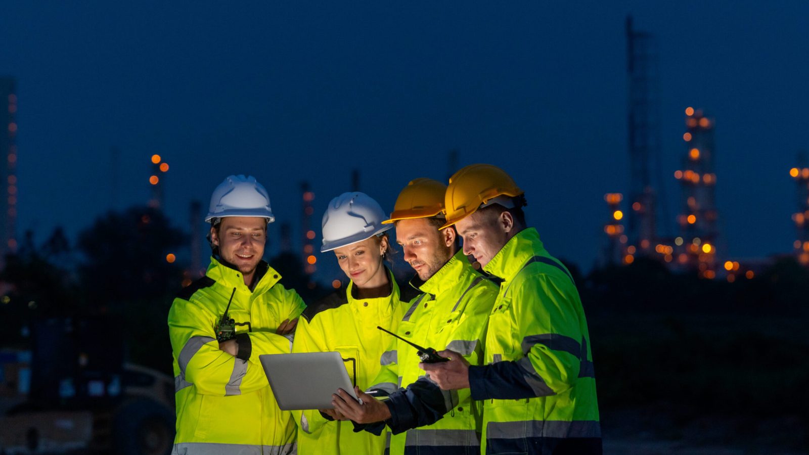 Group of petrochemical or gas workers or engineers use laptop to work in workplace area at night with light of factory in background.