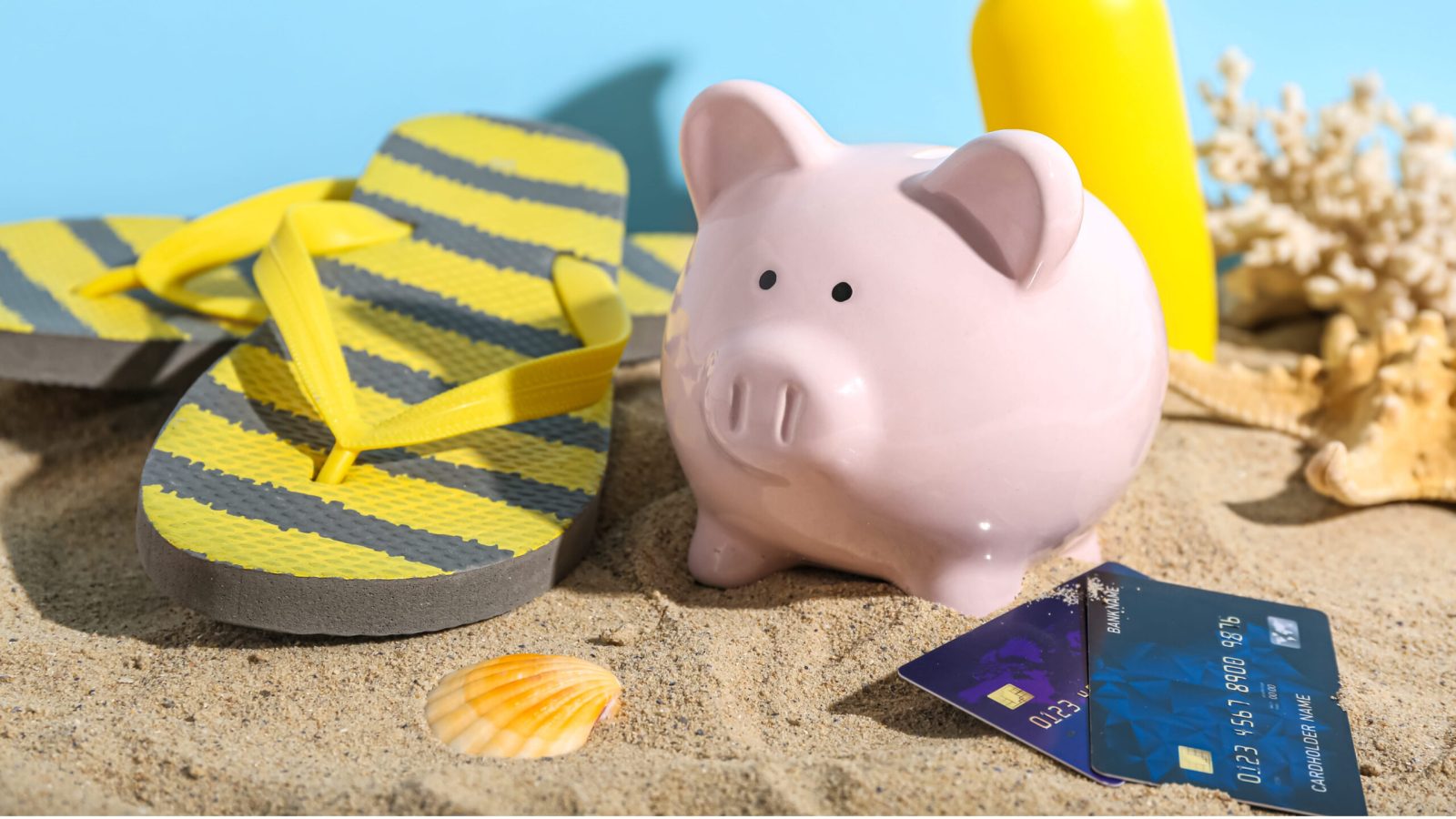 Piggy bank with beach accessories, sunscreen cream and credit cards on sand near blue wall. Concept of savings for travel